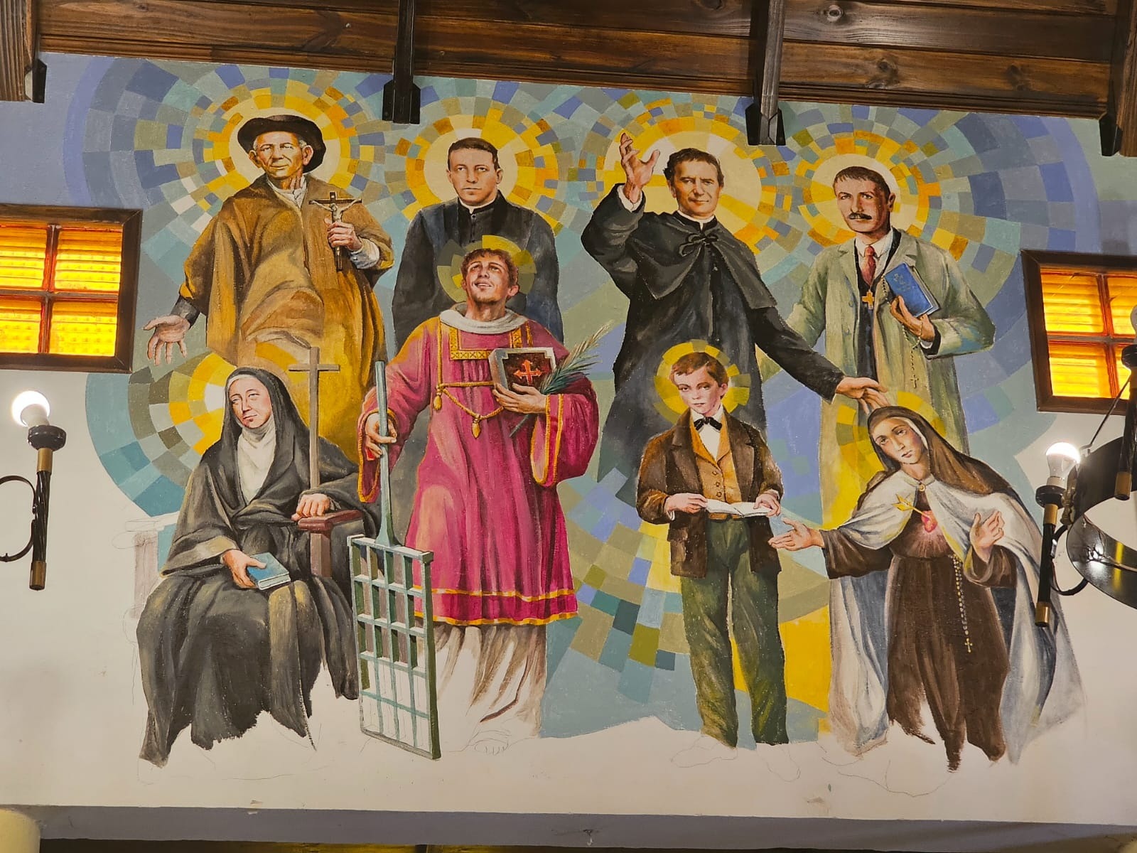 Mural de arte sacro realizado por Hugo Lazzarini que representa a santos y figuras emblemáticas de la Iglesia Católica, incluyendo al Cura Brochero, San Juan Bosco, San Lorenzo con su parrilla, Artémides Zatti, Ceferino Namuncurá, Santo Domingo Savio y Mama Antula. La obra destaca por sus características aureolas geométricas de colores vibrantes y un estilo de iconografía moderna.